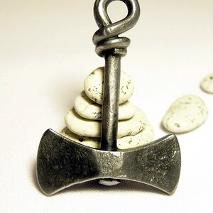 May include: A silver double-headed axe pendant with a knotted handle, resting on a stack of white stones.