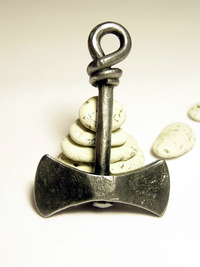 May include: A silver double-headed axe pendant with a knotted handle, resting on a stack of white stones.