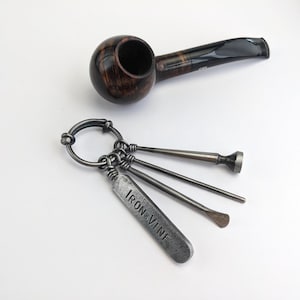 May include: A dark brown wooden tobacco pipe with a black stem positioned above a metal keyring holding three silver-toned tools, including a flat rectangular tag stamped with the text IRON VINE and two slender implements with rounded ends.