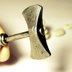 May include: A small, silver-colored, double-headed axe with a wooden handle. The axe is held in a hand, and the background is blurred.