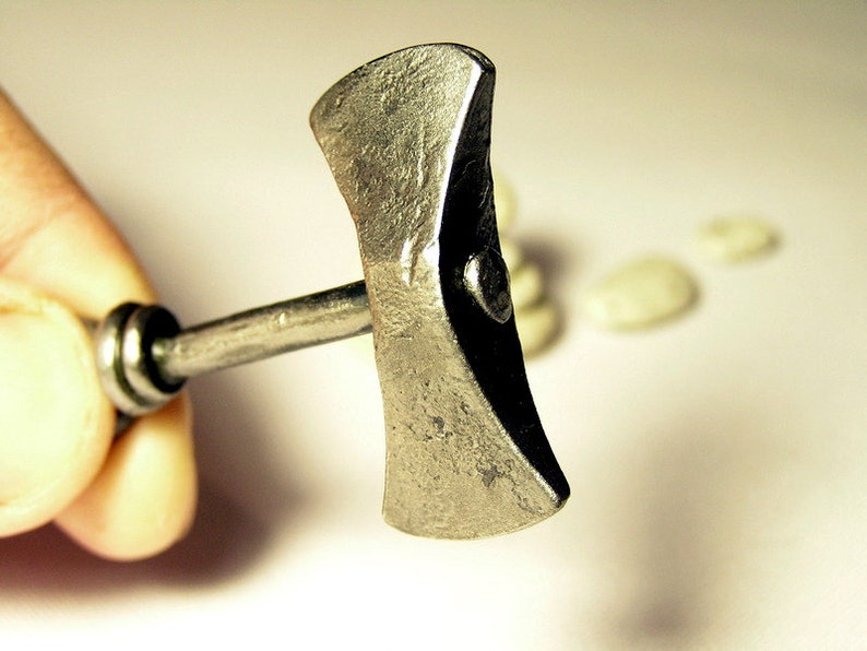 May include: A small, silver-colored, double-headed axe with a wooden handle. The axe is held in a hand, and the background is blurred.