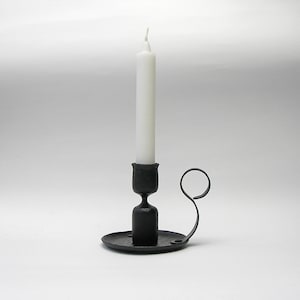 Peut inclure: Un bougeoir en métal noir avec une seule bougie blanche. Le bougeoir a un bras incurvé et une base ronde.