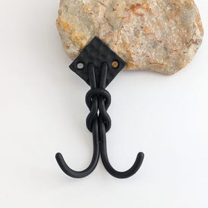 Puede incluir: Un gancho de hierro forjado a mano, de color negro, con una parte superior en forma de diamante y dos ganchos curvos. El gancho está sujeto a una piedra de color claro. El gancho está diseñado para colgar objetos.