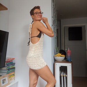 May include: A woman wearing a white crochet dress with a low back and a black bikini top underneath. She is standing in a room with a white wall and a wooden floor.