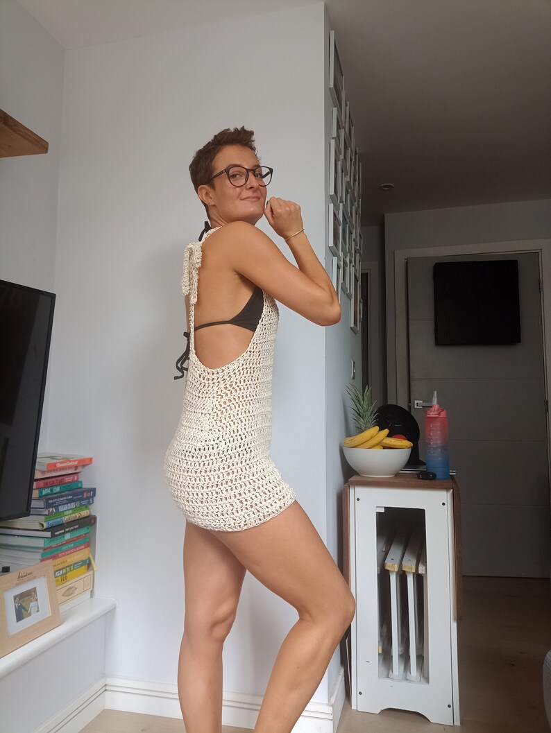 May include: A woman wearing a white crochet dress with a low back and a black bikini top underneath. She is standing in a room with a white wall and a wooden floor.