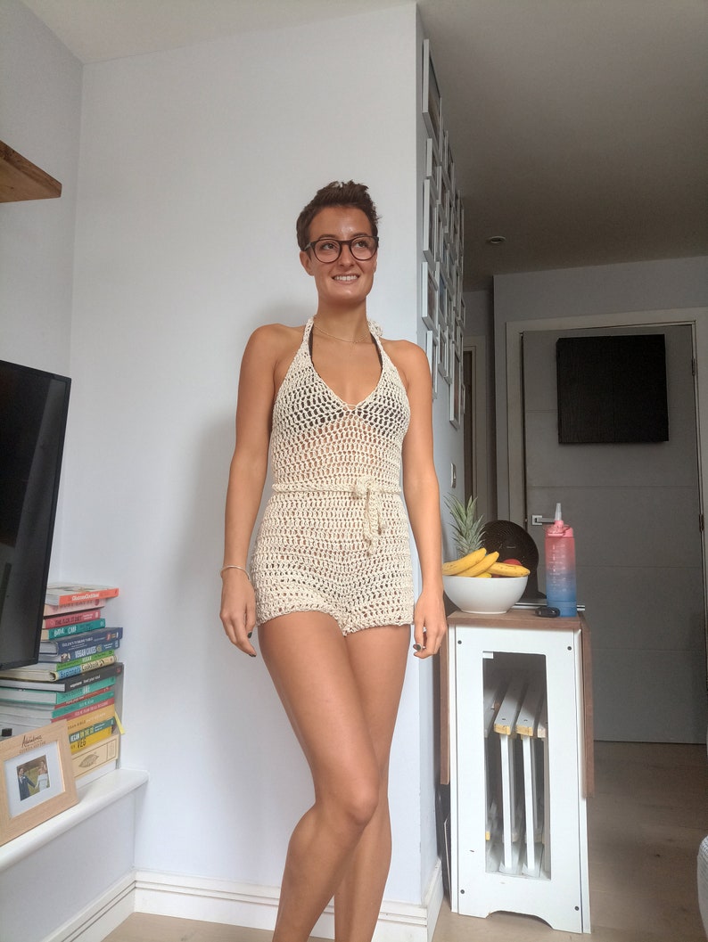 May include: A woman wearing a white crochet halter top and matching shorts. She is standing in a room with a white wall and a wooden table with fruit on it.