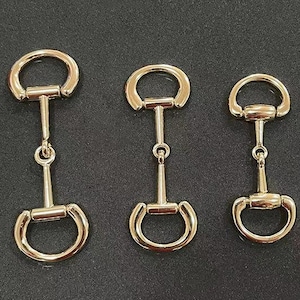 May include: Three gold-toned metal snaffle bit buckles of different sizes. The buckles are all the same design, with a round loop on each end and a straight bar in the middle.
