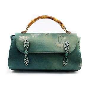 Puede incluir: Bolso de mano verde azulado con asa de bamb&uacute; y cierres plateados en forma de hoja. El bolso presenta un estampado floral en la solapa y una forma rectangular. El asa es de bamb&uacute; y est&aacute; sujeta con herrajes met&aacute;licos dorados.
