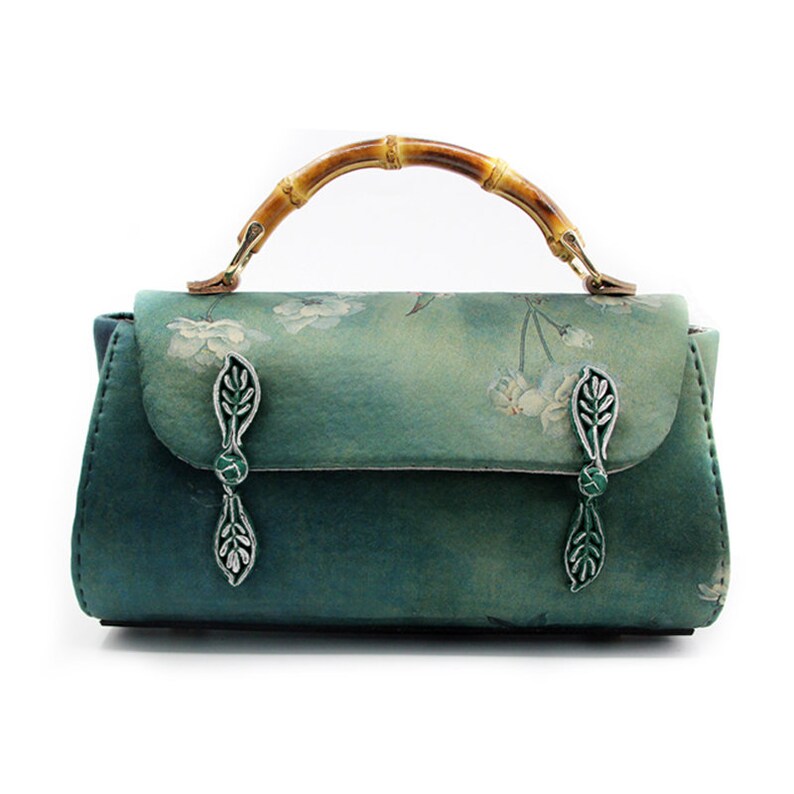Puede incluir: Bolso de mano verde azulado con asa de bamb&uacute; y cierres plateados en forma de hoja. El bolso presenta un estampado floral en la solapa y una forma rectangular. El asa es de bamb&uacute; y est&aacute; sujeta con herrajes met&aacute;licos dorados.