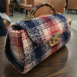 Puede incluir: Bolso rectangular con estampado de cuadros rojos, blancos y azul marino. Cuenta con un asa de bamb&uacute;, herrajes dorados y cierre de broche. El bolso est&aacute; sobre una superficie de madera.