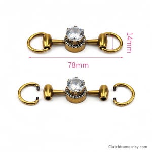 May include: Two antique gold-colored metal clasps with clear crystal center stones. Each clasp is 78mm long and 14mm high. The clasps have a ring on each end and are likely used for bags or other accessories.