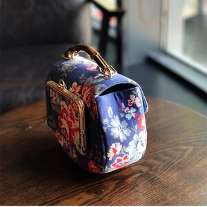 Puede incluir: Un bolso azul floral con asa de bamb&uacute; y herrajes dorados. El bolso presenta un cierre de broche y un peque&ntilde;o bolsillo en la parte delantera. El estampado floral incluye flores rojas, rosas y blancas.