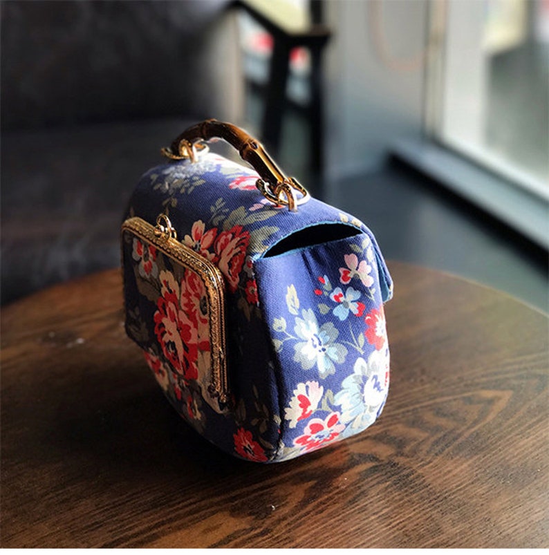 Puede incluir: Un bolso azul floral con asa de bamb&uacute; y herrajes dorados. El bolso presenta un cierre de broche y un peque&ntilde;o bolsillo en la parte delantera. El estampado floral incluye flores rojas, rosas y blancas.