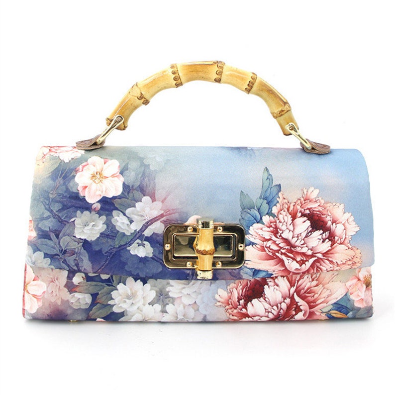 Puede incluir: Bolso de mano rectangular con asa de bamb&uacute; y herrajes dorados. El bolso presenta un estampado floral con flores rosas y blancas sobre un fondo azul. El asa es de bamb&uacute; curvado.