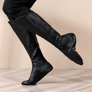 A person wearing black leather boots is standing on a wooden floor.