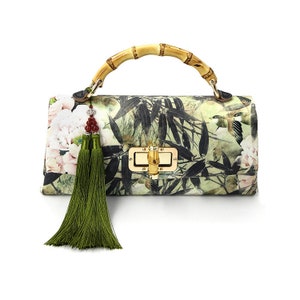 Puede incluir: Un bolso rectangular con asa de bamb&uacute; y cierre dorado. El bolso presenta un estampado floral y de p&aacute;jaros en tonos verdes, blancos y negros. Una larga borla verde cuelga del lateral, a&ntilde;adiendo un elemento decorativo.