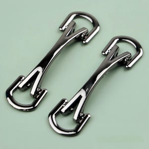 May include: Two shiny, gunmetal-colored metal clasps. Each clasp has a curved top and a stylized, angular design. The clasps are positioned on a light green background, showcasing their reflective surfaces and intricate details.