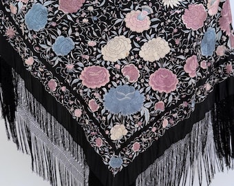 May include: A large black triangular shawl featuring intricate floral embroidery in shades of dusty pink, light blue, and cream. The fabric is adorned with dense botanical patterns and finished with long, black knotted fringe along the edges.