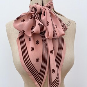 May include: A pink silk scarf featuring a brown geometric pattern of circles and stripes. The scarf is tied in a bow around a mannequin's neck, highlighting its design. This accessory adds a touch of vintage elegance.