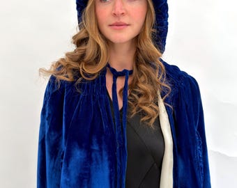 May include: A deep royal blue velvet hooded cape featuring a gathered neckline with matching velvet tie closures. The cape is lined with a smooth, light-colored fabric and drapes over the shoulders, providing full coverage with a soft texture.