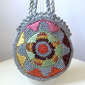 May include: A round, silver-coloured crocheted handbag featuring two braided handles. The front surface displays a circular, multi-coloured embroidered pattern in shades of pink, yellow, light green, dark red, and orange against a silver base.