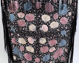 May include: A large black rectangular shawl featuring intricate floral embroidery in shades of dusty pink, light blue, and cream. The fabric is adorned with dense botanical patterns and finished with long black fringe along the edges.