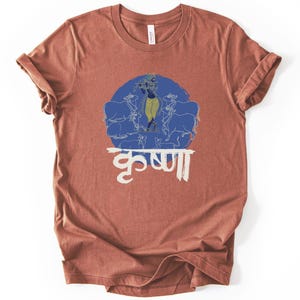 Puede incluir: Camiseta de color óxido con un diseño circular azul que representa una figura tocando la flauta, rodeada de vacas. La palabra "Krishna" está escrita en escritura blanca debajo. Las mangas están enrolladas.
