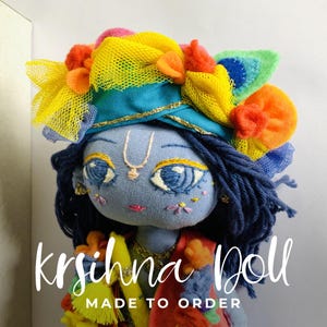 May include: A fabric doll with light blue skin, dark blue yarn hair, and embroidered facial features. The doll wears a vibrant headpiece featuring yellow netting, orange pom-poms, and a green peacock feather. Text reads: krishna Doll MADE TO ORDER.