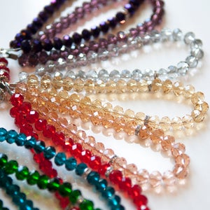 May include: Close-up of several beaded bracelets in various colors, including green, blue, red, gold, silver, and purple. The beads are faceted and appear to be made of glass or crystal, reflecting light. The bracelets are arranged on a white surface.