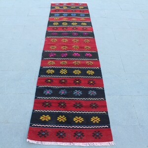 May include: A long, narrow red and black rug with a geometric pattern. The rug has a fringe on both ends.
