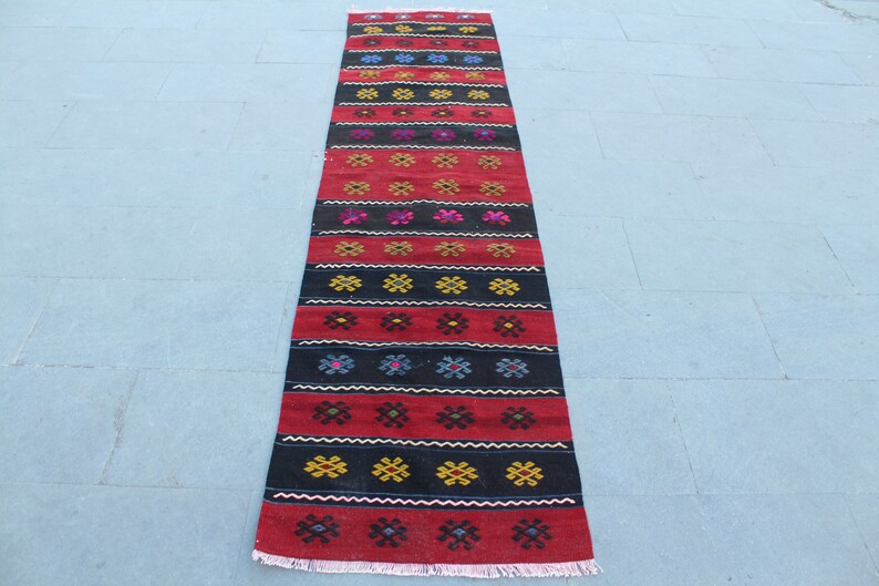 May include: A long, narrow red and black rug with a geometric pattern. The rug has a fringe on both ends.