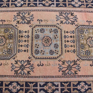 May include: A vintage rug with a pink background and intricate geometric patterns in shades of blue, brown, and beige. The rug features a central design with three repeating motifs.