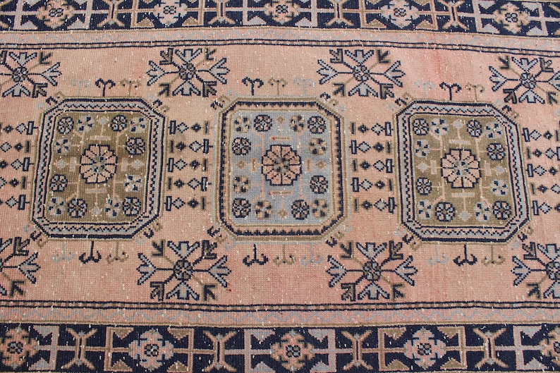 May include: A vintage rug with a pink background and intricate geometric patterns in shades of blue, brown, and beige. The rug features a central design with three repeating motifs.