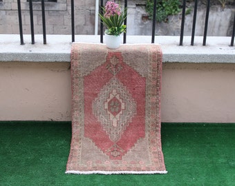 May include: A pink and beige vintage rug with a geometric pattern. The rug is laying on a green surface and a white pot with pink flowers is on a ledge behind the rug.