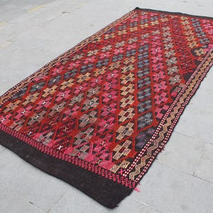 May include: A traditional Turkish rug with a geometric pattern in shades of red, brown, and black. The rug has a dark brown background with intricate details woven into the design.