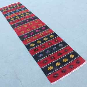 May include: A long, narrow red and black rug with a geometric pattern. The rug has a fringe on both ends.
