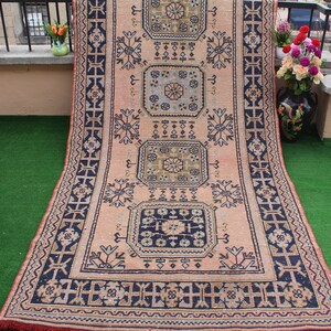 May include: A vintage rug with a pink background and intricate blue and green geometric patterns. The rug has a fringed edge and is laid out on a green surface.