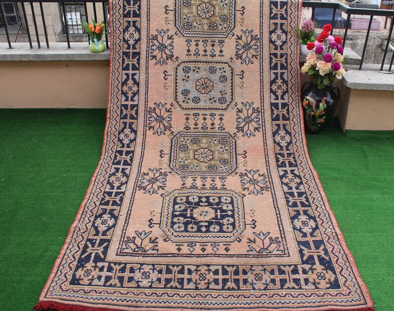 May include: A vintage rug with a pink background and intricate blue and green geometric patterns. The rug has a fringed edge and is laid out on a green surface.