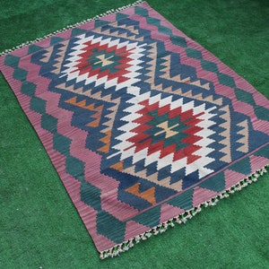 Puede incluir: Una alfombra colorida con un patr&oacute;n geom&eacute;trico en tonos de rojo, azul, verde, blanco y marr&oacute;n. La alfombra tiene un borde de flecos y est&aacute; colocada sobre una superficie verde.
