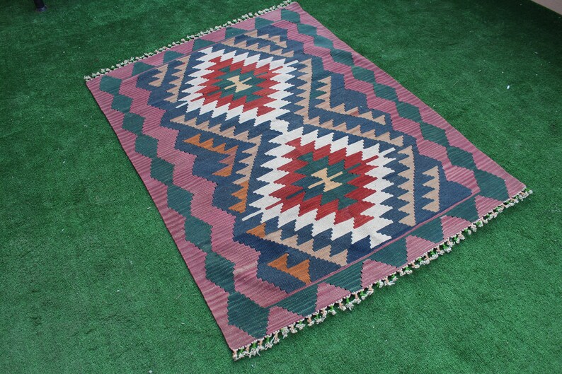 Puede incluir: Una alfombra colorida con un patr&oacute;n geom&eacute;trico en tonos de rojo, azul, verde, blanco y marr&oacute;n. La alfombra tiene un borde de flecos y est&aacute; colocada sobre una superficie verde.