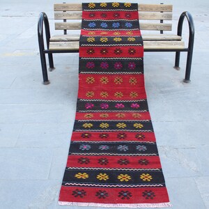 May include: A long, narrow red and black rug with a geometric pattern of flowers and squares. The rug is laying on a bench.