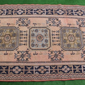 May include: A vintage rug with a pink background and intricate geometric patterns in shades of blue, green, and brown. The rug has a border with a repeating pattern of crosses.
