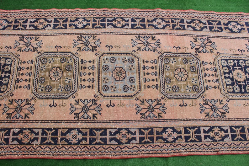 May include: A vintage rug with a pink background and intricate geometric patterns in shades of blue, green, and brown. The rug has a border with a repeating pattern of crosses.
