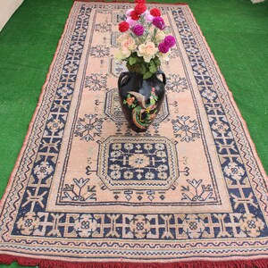 May include: A vintage rug with a floral pattern in shades of pink, blue, and white. The rug has a fringe border and is decorated with a black vase filled with pink and white flowers.