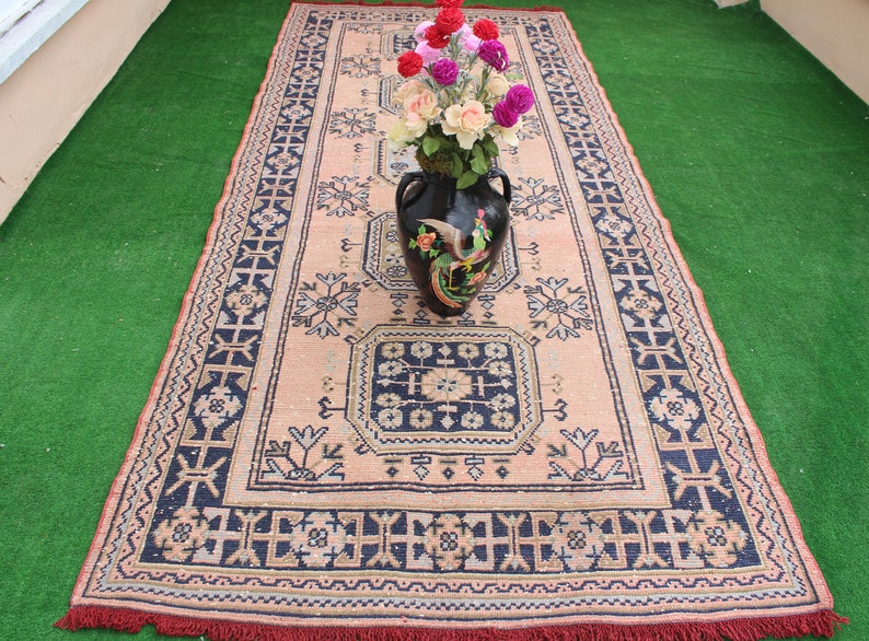 May include: A vintage rug with a floral pattern in shades of pink, blue, and white. The rug has a fringe border and is decorated with a black vase filled with pink and white flowers.