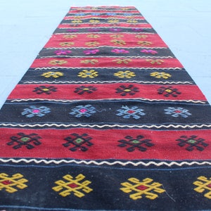 May include: A long, narrow rug with a traditional geometric pattern in shades of red, black, white, blue, and yellow. The rug is woven with intricate details and has a rustic, handmade look.
