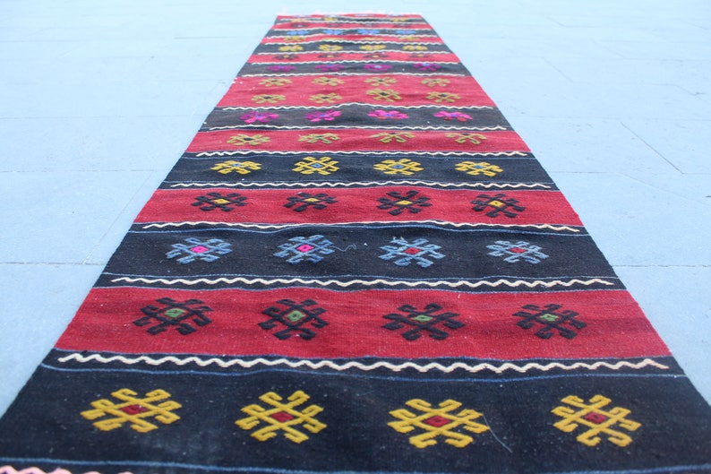 May include: A long, narrow rug with a traditional geometric pattern in shades of red, black, white, blue, and yellow. The rug is woven with intricate details and has a rustic, handmade look.