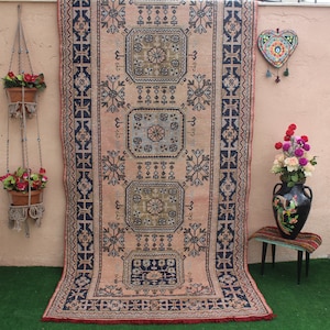 May include: A vintage rug with a pink background and intricate blue and black geometric patterns. The rug has a fringe border and is hanging on a wall.