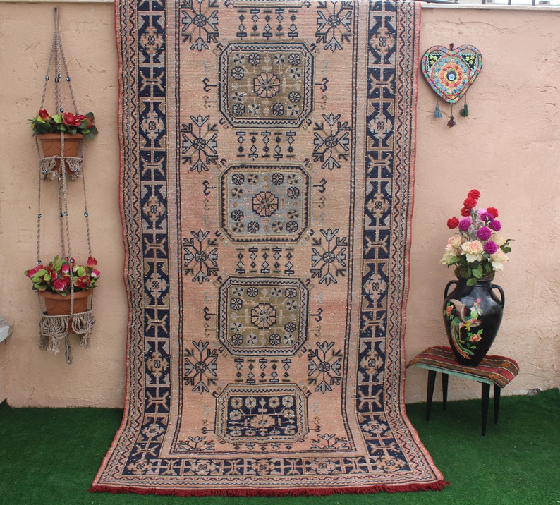 May include: A vintage rug with a pink background and intricate blue and black geometric patterns. The rug has a fringe border and is hanging on a wall.