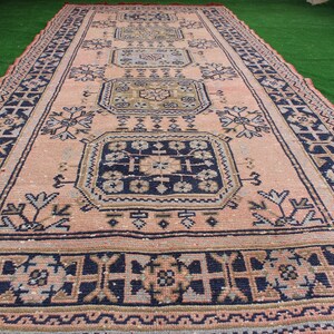May include: A vintage rug with a pink background and intricate blue, brown, and white patterns. The rug has a geometric design with repeating motifs.
